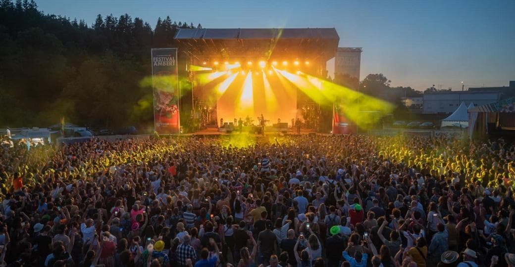 Foule de spectateurs devant une grande scène extérieure éclairée de faisceaux orangés, au crépuscule